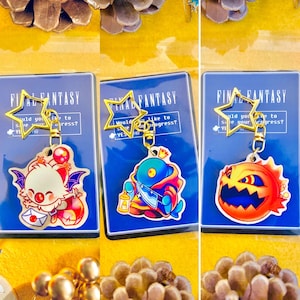 Könnte beinhalten: Drei Final Fantasy Schlüsselanhänger mit einem blauen und roten Charakter, einem weißen und roten Charakter und einem roten und orangefarbenen Charakter. Jeder Anhänger hat einen goldenen sternförmigen Ring und den Text "Final Fantasy" auf blauem Hintergrund.