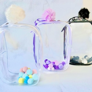 Op de afbeelding: Drie doorzichtige, rechthoekige cosmetische tassen met kleurrijke pompons erin. Elke tas heeft een andere gekleurde pompon bovenop: wit, paars en zwart. De tassen zijn gevuld met kleine, kleurrijke pompons.