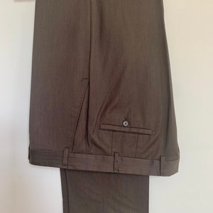 Può includere: Un paio di pantaloni da abito marroni appesi a un'appendiabiti bianca. I pantaloni hanno un sottile motivo a righe sottili e passanti per la cintura.