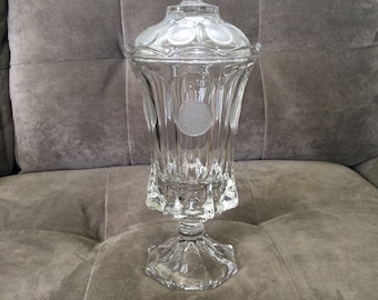 May include: A clear glass pedestal with a lid, featuring a decorative etched design of circles. The pedestal is on a brown couch.