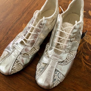 Peut inclure: Une paire de chaussures à lacets argentées et blanches avec un motif floral. Les chaussures ont un éclat métallique et des lacets blancs. Le design comprend une combinaison de matériaux argentés et blancs, créant une esthétique vintage.