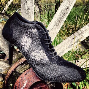 May include: Black suede heeled shoe with a lace-up closure and a textured pattern. The shoe is sitting on a wooden surface.