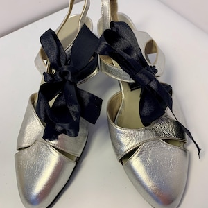 Op de afbeelding: Een paar zilveren slingback-hakken met een zwarte satijnen strik. De schoenen hebben een spitse neus en een uitgesneden ontwerp aan de zijkanten. De hakken zijn ongeveer 7,6 cm hoog.