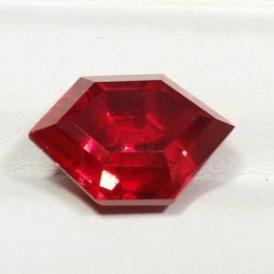 May include: A vibrant, deep red gemstone cut into a unique hexagonal shape with multiple facets that reflect light. The stone displays a brilliant, saturated crimson hue and a polished, smooth surface, showcasing its geometric, multi-sided structure.