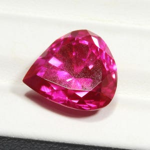 May include: A pear-shaped, faceted gemstone with a vibrant, deep pinkish-red color. The stone features a brilliant cut with multiple triangular and kite-shaped facets that reflect light, creating a sparkling appearance across its surface.