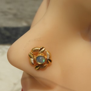 May include: A gold-coloured nose stud with a central, round, opalescent stone. The stud is surrounded by a decorative, floral-inspired design. The jewellery is set against a light skin tone.