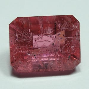 May include: A faceted, rectangular-cut gemstone with a vibrant pink hue. The gem has a slightly translucent appearance, with internal inclusions and subtle variations in color. The edges are precisely cut, and the overall shape is symmetrical.