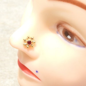 Könnte beinhalten: Ein goldener Nasenring mit einem roten Edelstein in der Mitte. Der Ring hat die Form einer Blume mit acht Blütenblättern.