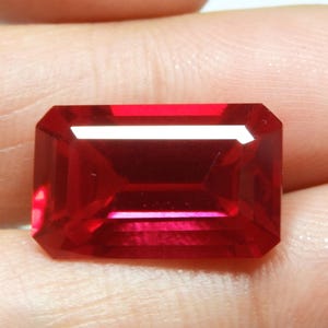 May include: A faceted, rectangular-cut, deep red gemstone. The gem has a brilliant, reflective surface with sharp edges and a polished appearance. The color is a rich, saturated red, with internal facets visible.
