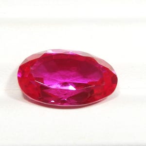 May include: A vibrant, oval-cut gemstone with a deep, saturated red color. The stone features numerous triangular and kite-shaped facets across its surface, which create a brilliant, reflective appearance and highlight its translucent quality.
