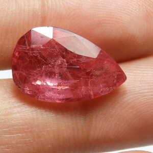 May include: A faceted, pear-shaped gemstone with a vibrant pink hue. The gem displays internal inclusions and a polished surface, reflecting light. The stone is held between fingers, showcasing its size and color.