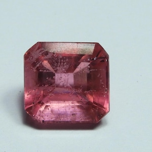 May include: A faceted, square-cut pink gemstone with a slightly translucent appearance. The gem has a vibrant pink hue with subtle internal inclusions, set against a white background. The gemstone's cut showcases its facets.