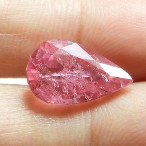 May include: A faceted, teardrop-shaped gemstone in shades of pink. The gem has a complex internal structure with visible inclusions and a high degree of transparency. The cut of the stone creates multiple reflective surfaces.