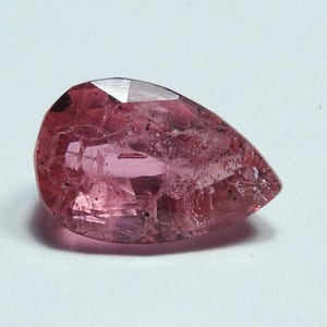 May include: A faceted, pear-shaped gemstone with a pink hue. The gem has a translucent quality, with internal inclusions and a polished surface. The cut reveals multiple facets, enhancing its brilliance. The gemstone is likely a tourmaline or similar precious stone.