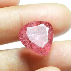 May include: A faceted, heart-shaped gemstone with a pink hue. The gem has a central, lighter-colored facet and a slightly speckled appearance. The stone is held between two fingers against a white background.