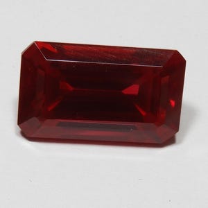 Puede incluir: Una gema roja oscura tallada en forma rectangular. La gema tiene una superficie pulida y bordes afilados, reflejando la luz. La piedra, similar a un rubí, está sobre un fondo blanco. Dimensiones: aproximadamente 2 cm x 1 cm.