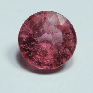May include: A faceted, round, pink gemstone with internal inclusions. The gem is a deep rose color, with a polished surface that reflects light. The gemstone is set against a white background.