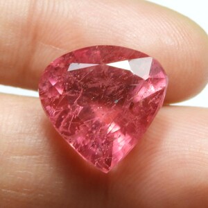 May include: A faceted, pear-shaped gemstone with a vibrant pink hue. The gem displays internal inclusions and a high degree of transparency, reflecting light across its many facets. The gemstone is held between fingers, showcasing its size and color.