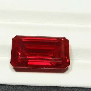 May include: A faceted, rectangular-cut, deep red gemstone. The gem is set against a white background, showcasing its clarity and vibrant color. The cut of the stone allows light to reflect, creating a brilliant display.