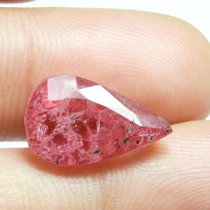 May include: A faceted, teardrop-shaped gemstone with a vibrant pink hue. The gem has internal inclusions and dark specks, and is held between two fingers. The cut of the stone creates multiple reflective surfaces.