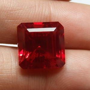 May include: A faceted, rectangular-cut, deep red gemstone. The gem is held between fingers, showcasing its brilliant, reflective surfaces. The gem's color is a rich, saturated red, with sharp, clean edges.