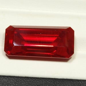 May include: A faceted, rectangular-cut, deep red gemstone. The gem has a polished surface with sharp edges and a brilliant shine. The stone is set against a white background.