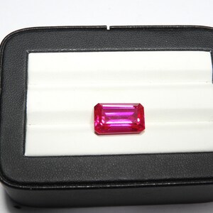 May include: A rectangular, vibrant pink gemstone is displayed in a black jewelry box with a white interior. The gemstone is the focal point, showcasing its polished facets and rich color. The box provides a contrasting backdrop, highlighting the gem's brilliance.