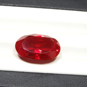 May include: A faceted, oval-shaped, deep red gemstone. The gem is set against a white background within a black-edged display case. The stone's polished surfaces reflect light, showcasing its clarity and color.