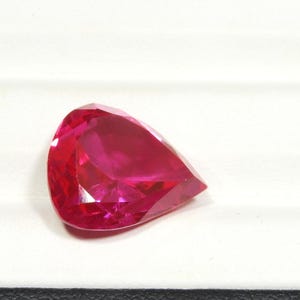 May include: A pear-shaped gemstone with a vibrant, deep red color and multiple facets that reflect light. The stone features a pointed end and a rounded base, showcasing a clear, transparent quality with sharp, geometric edges throughout its surface.