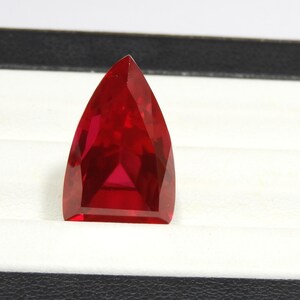 Puede incluir: Una piedra preciosa facetada en forma de escudo con un color rojo intenso y vibrante. La piedra presenta una parte superior puntiaguda y una base plana, con múltiples facetas geométricas que reflejan la luz en toda su superficie.
