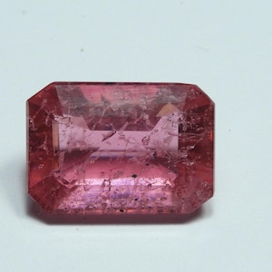 May include: A faceted, rectangular gemstone with a pink hue. The gem has a slightly irregular surface with internal fractures, and is set against a white background. The gemstone's cut features multiple facets, enhancing its brilliance.