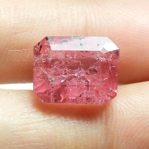 May include: A faceted, rectangular pink gemstone with a slightly irregular surface. The gem has a light pink hue with darker inclusions and is held in a person's hand. The cut is an emerald cut, with a flat top and beveled edges.