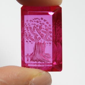 May include: A rectangular, translucent, hot pink gemstone featuring an intaglio carving of a tree with a textured trunk and rounded leaves. The stone has faceted edges and a polished surface, displaying a vibrant, saturated magenta color throughout.