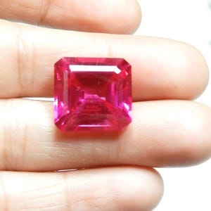 May include: A radiant, rectangular-cut, pink gemstone. The gem is held in a hand, highlighting its facets and internal brilliance. The stone's colour ranges from deep magenta to lighter pink hues, reflecting light.