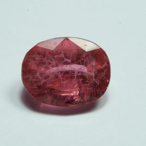 May include: A faceted, oval-shaped gemstone with a deep pink hue. The gem has internal inclusions and a polished surface, reflecting light. The stone's shape is symmetrical, with a flat top and a rounded bottom. The gem is set against a white background.