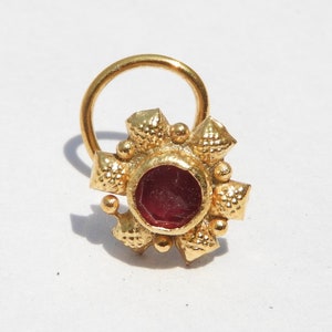 May include: A gold nose ring with a red gemstone in the centre. The ring has a decorative design with small, raised dots and points.