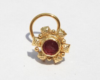 May include: A gold nose ring with a red gemstone in the center. The ring has a decorative design with small, raised dots and points.