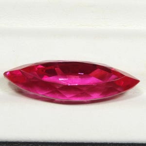 May include: A marquise-cut gemstone with a vibrant, saturated fuchsia-pink color. The stone features a faceted surface that reflects light, creating a brilliant appearance with sharp, pointed ends and a wider, rounded center section.