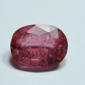 May include: A faceted, oval-shaped gemstone with a pink hue. The gem has a polished surface with internal inclusions and a reflective, crystalline appearance. The gem is set against a white background.