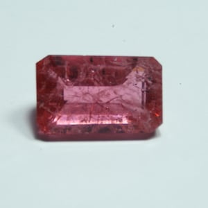 May include: A rectangular, faceted gemstone with a pinkish-red hue. The gem has a clear, crystalline appearance with internal inclusions. The cut is an emerald cut, with a flat top and a series of rectangular facets. The gemstone is set against a white background.
