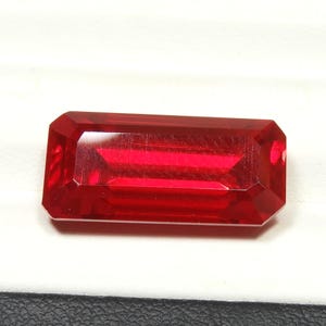 May include: A faceted, rectangular-cut, deep red gemstone. The gem has a polished surface with sharp edges and a high degree of transparency, reflecting light. The gemstone is set against a white and black background.