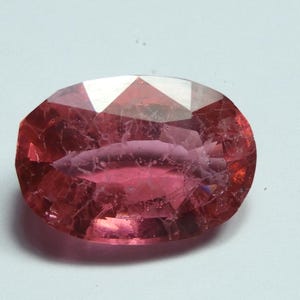 May include: A faceted, oval-shaped gemstone with a pinkish-red hue. The gem has a complex internal structure with visible inclusions and a polished surface. The light reflects off the facets, creating a sparkling effect. The gemstone is set against a white background.