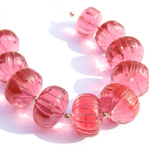 Puede incluir: Un collar de cuentas de vidrio rosa con un diseño acanalado. Las cuentas ovaladas están ensartadas en un cordón fino y transparente. Las cuentas rosas translúcidas se muestran sobre un fondo blanco. Cada cuenta mide aproximadamente 1,5 cm de diámetro.