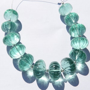 Puede incluir: Un collar de cuentas de vidrio de color verde azulado claro. Cada cuenta tiene forma de calabaza acanalada y está ensartada con un espaciador plateado. Las cuentas están sobre un fondo blanco, proyectando sombras.