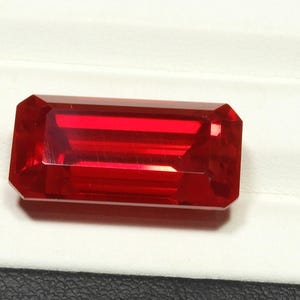 Peut inclure: Une pierre précieuse rouge vif taillée en forme de baguette rectangulaire avec des bords facettés. La pierre présente une teinte cramoisie profonde et saturée ainsi qu'une surface polie et réfléchissante qui capte la lumière.