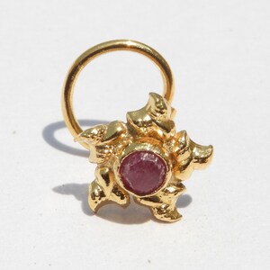 May include: A gold nose ring with a floral design and a central, round, red gemstone. The ring has a circular loop for insertion and a decorative, textured flower shape around the gemstone.