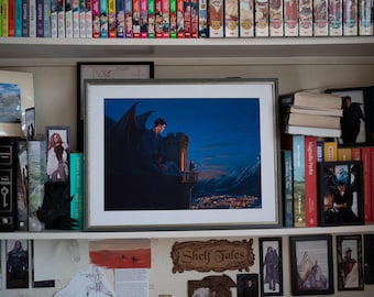 May include: A framed print of a fantasy scene with a dark-winged figure on a castle tower overlooking a city at dusk. The print is on a shelf with books and other framed art. The shelf is filled with various books and decorative items.