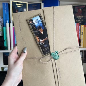 May include: A rectangular brown paper package wrapped in twine with a green wax seal. A vertical bookmark featuring a male figure and mountain landscape is tucked under the twine. Handwritten text reads To the stars who listen and Happy reading.