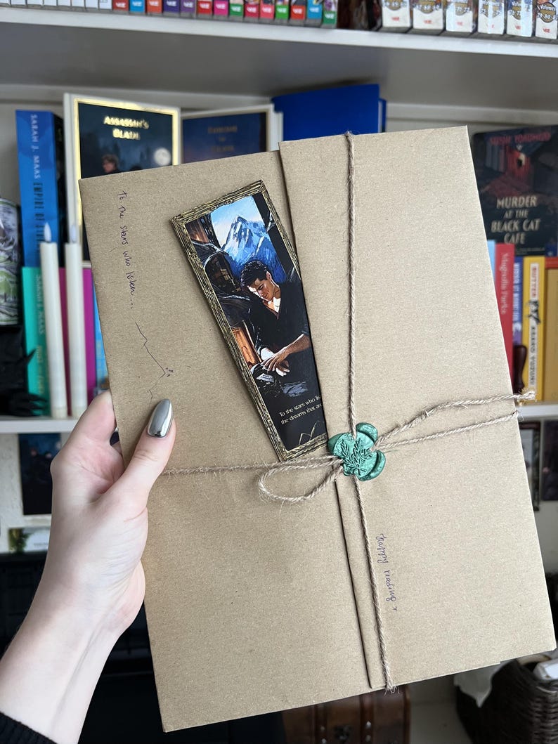 May include: A rectangular brown paper package wrapped in twine with a green wax seal. A vertical bookmark featuring a male figure and mountain landscape is tucked under the twine. Handwritten text reads To the stars who listen and Happy reading.