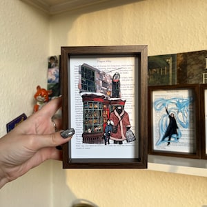 Może przedstawiać: Oprawione dzieło sztuki na papierze przedstawiające Ulicę Pokątną z serii Harry Potter. Grafika przedstawia szczegółową ilustrację sklepów i postaci na tle stron książki. Rama jest w kolorze ciemnobrązowym.